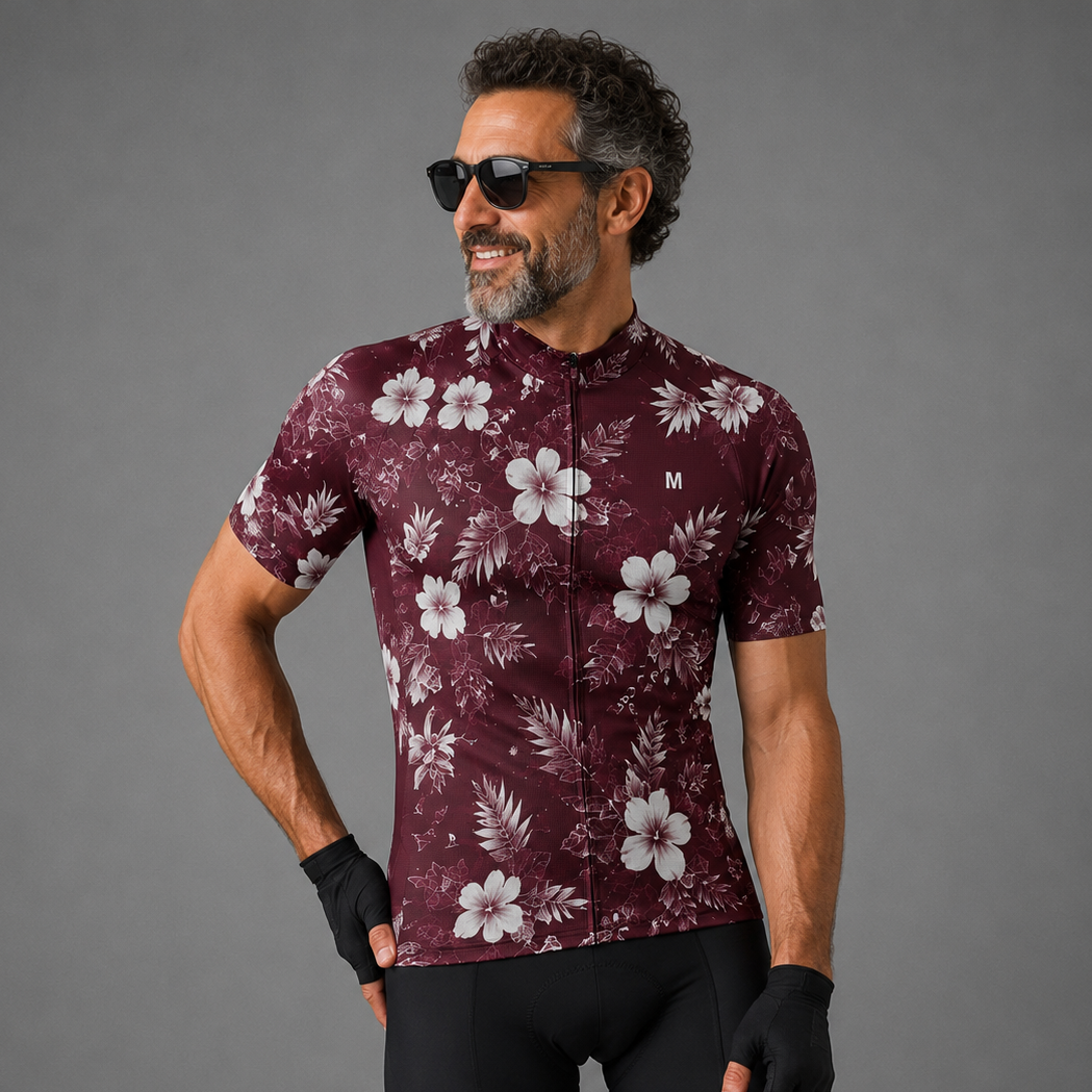 A man wearing a short-sleeve cycling jersey with a white floral print on a burgundy base, paired with black cycling shorts and fingerless gloves, posing against a solid gray studio background.