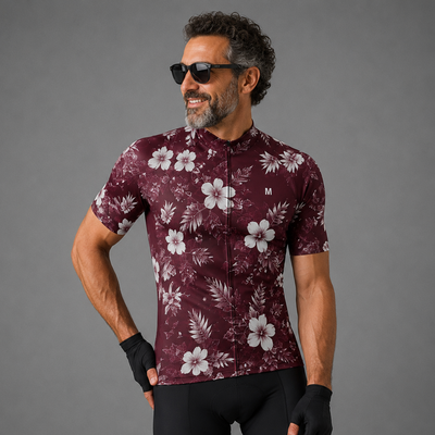 A man wearing a short-sleeve cycling jersey with a white floral print on a burgundy base, paired with black cycling shorts and fingerless gloves, posing against a solid gray studio background.