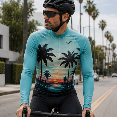 A man wearing a turquoise long-sleeve cycling jersey with a tropical sunset and palm tree print, black shorts, and a black helmet rides a road bike on a palm-lined street.
