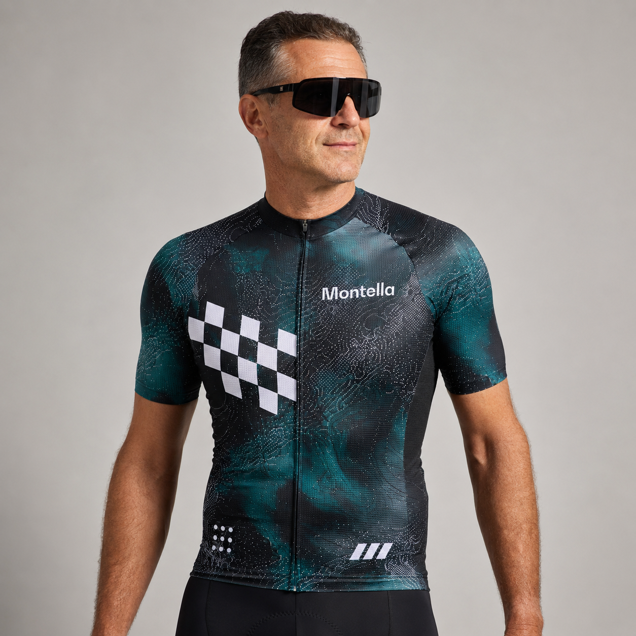 A man wearing a short-sleeve Montella cycling jersey with a dark teal and black abstract pattern, a white checkered graphic on the chest, and black sunglasses, photographed against a neutral gray studio background.