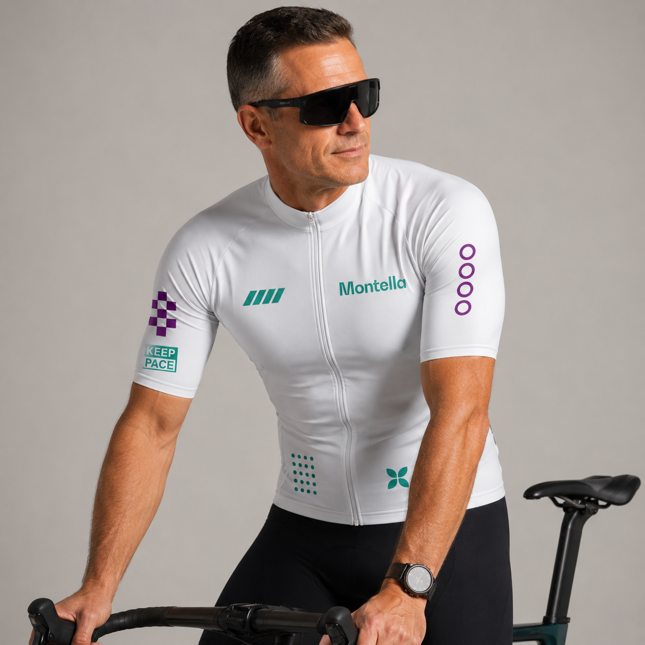 A man wearing a white Montella cycling jersey with green and purple sponsor logos, black cycling shorts, and black sunglasses, riding a bicycle against a neutral gray background.