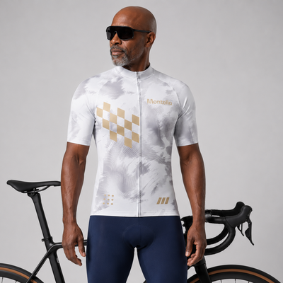A man wearing a white and gray patterned cycling jersey with gold accents and navy blue cycling shorts, standing in front of a black road bicycle against a light gray studio background.